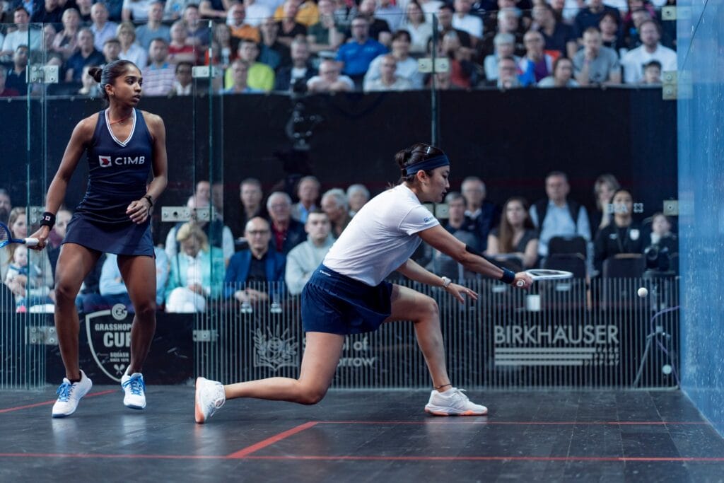 Satomi Watanabe spielt einen Ball; Sivasangari Subramaniam steht hinter ihr auf dem Glascourt.